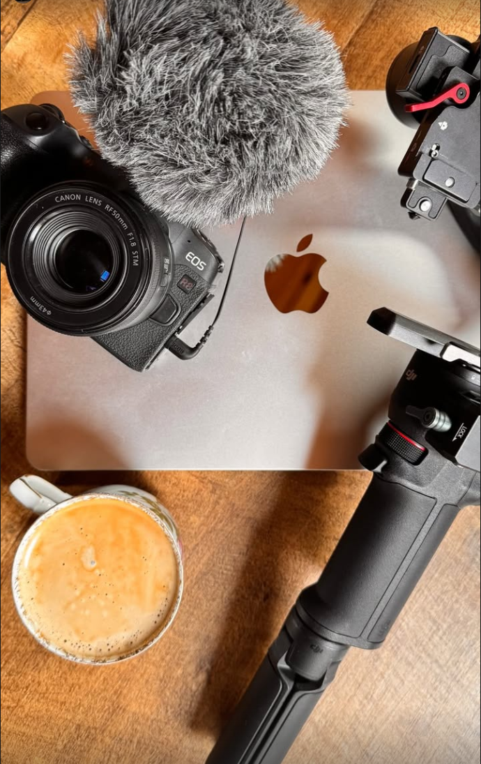 Biurko z MacBookiem, kubkiem kawy, aparatem Canon z obiektywem 50 mm, mikrofonem nakamerowym i gimbalem DJI.